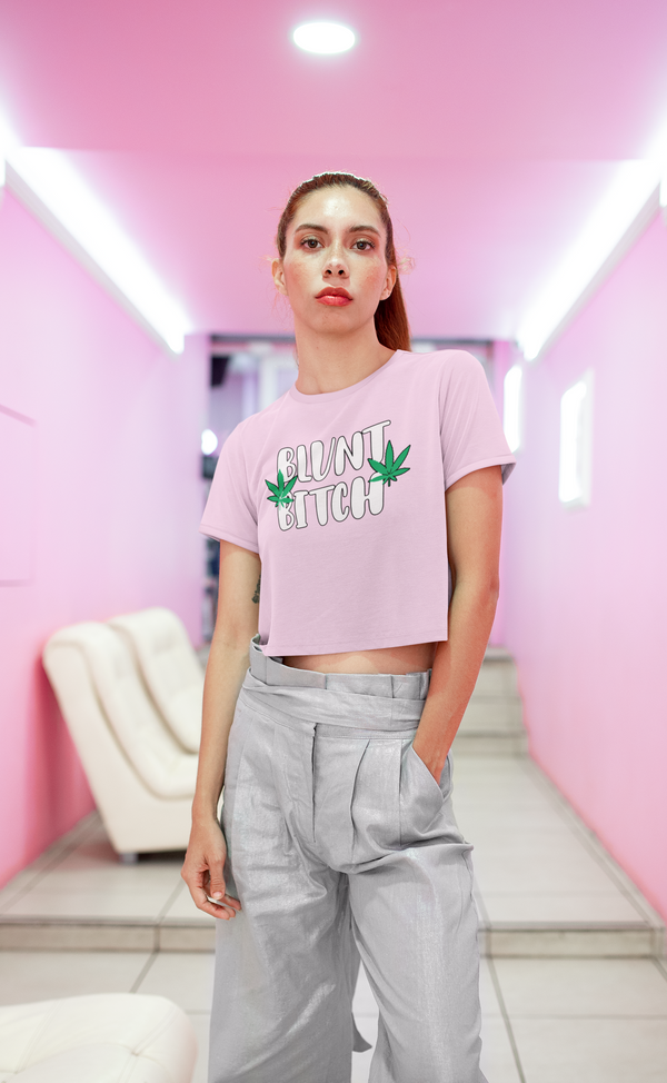 Casual fashion model posing in neon-lit hallway. White crop top with bold green logo design. Stylish gray pleated trousers. Eye-catching makeup and confident expression.